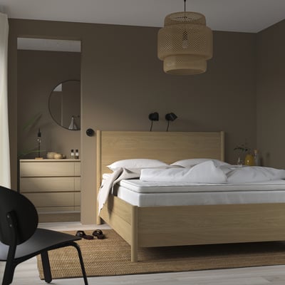 Ett mysigt sovrum, en träsäng med vita sängkläder, en vävd taklampa och en modern byrå med en rund spegel. En svart stol är placerad nära sängen.
