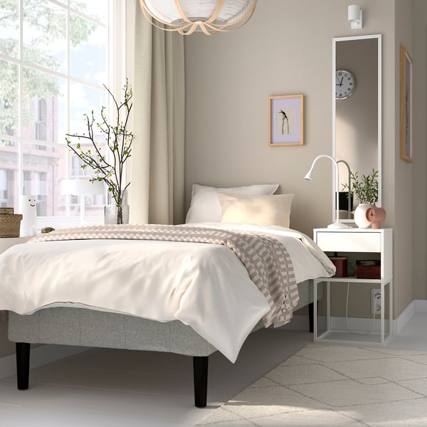 Sovrum med sängen RENFJÄLLET , vit, tyg, rektangulär, minimalistisk design. Sängen är större än nattduksbordet, spegeln och fönstret.