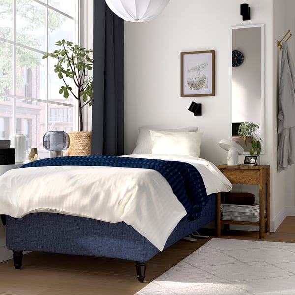 Modern vit säng, blå accenter, sidobord med lampa och växt, stora fönster, spegel, minimalistisk inredning.