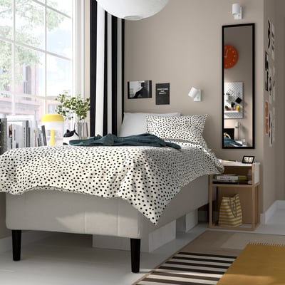 Modernt sovrum med sängen RISHÖJDEN , prickigt uppslag, stort fönster, nattduksbord, gul lampa, växt, böcker, spegel, konst, randig matta.