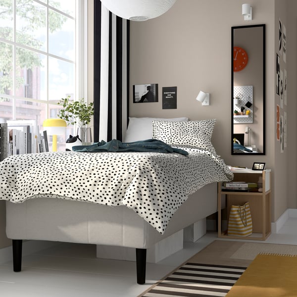 Modernt sovrum med grå säng, prickigt täcke, gul lampa, böcker och spegel.