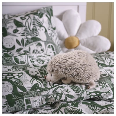 En grå SKOGSDUVA mjuk igelkottsleksak på en grön blommig bädd. Den är liten, rund och plyschig och placerad mellan stora kuddar.