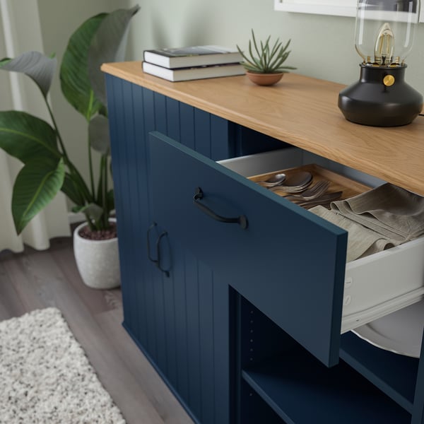 Öppen blå skåplåda med bestick, dukar, böcker, växt och lampa på träskiva.