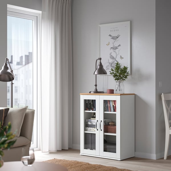 Modernt vardagsrum med vitt SKRUVBY vitrinskåp vid fönstret, ekaccenter, grå soffa, stol och minimalistisk inredning.