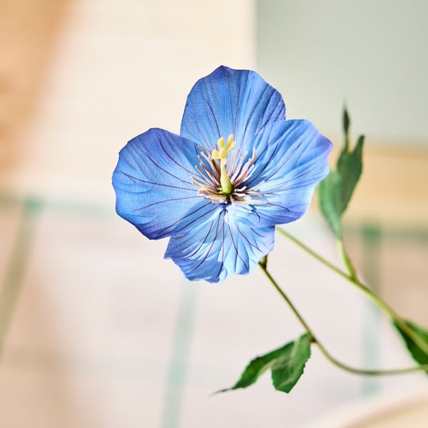 Klarblå konstgjord blomma, livfulla kronblad runt en gul mitt, fäst vid en grön stjälk med blad.