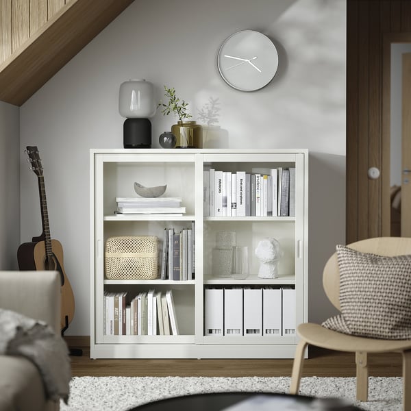 Minimalistiskt vardagsrum med vit bokhylla TONSTAD, med skjutdörrar, glasvaser, staplad inredning och väggklocka.