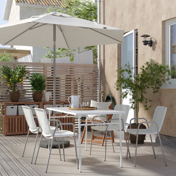 Modern uteservering med vita stolar och bord, stort beige paraply, krukväxter, trästaket och beige byggnad i bakgrunden.