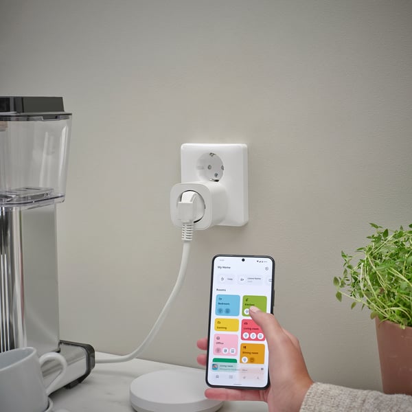 En person håller i en smartphone som styr en kontakt som är ansluten till en kaffebryggare via en smart hem-app, vilket demonstrerar fjärrhantering av enheter.
