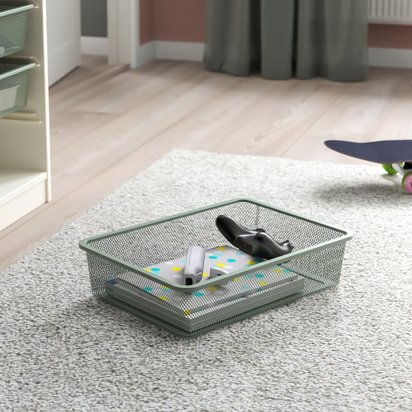 Förvaringskorg i mesh på matta, innehållande fjärrkontroll och elektroniska tillbehör, en del av TROFAST-serien för att organisera föremål.