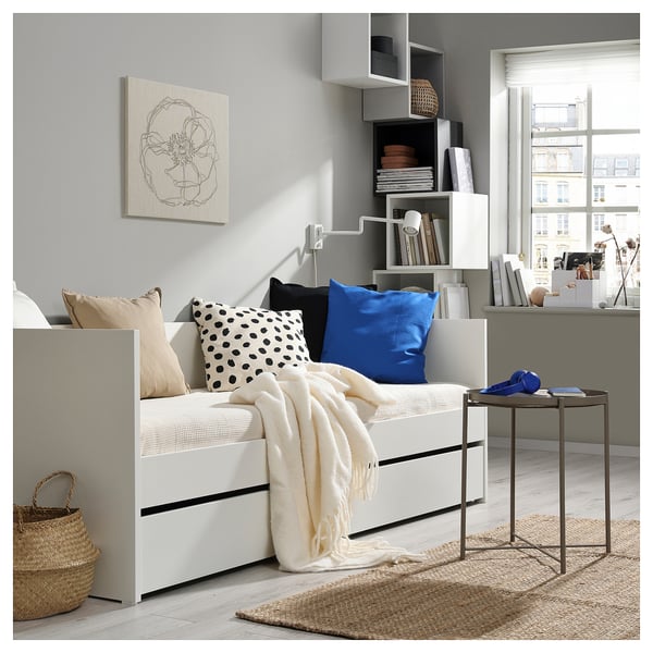 Mysigt sovrum med vit dagbädd, blå kudde, vägghyllor, konst, korg, litet bord.
