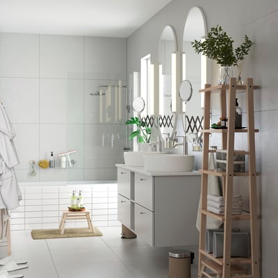 Modernt vitt badrum med dusch i glas, dubbla handfat, speglar och en hylla i ljust trä i stegstil fylld med handdukar och växter.