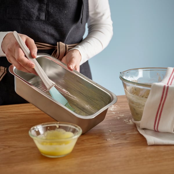 Personen smörjer brödformen med blå spatel och förbereder sig för att baka bröd. Plåt står på ett träbord med en skål med smält smör.