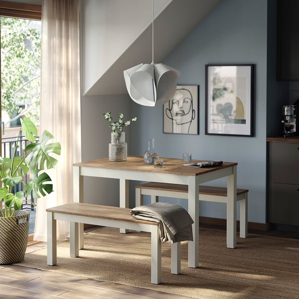 Modern dining spot with ÅLHULT light brown bench, grey throw, vase of white flowers, and abstract art on light blue wall.