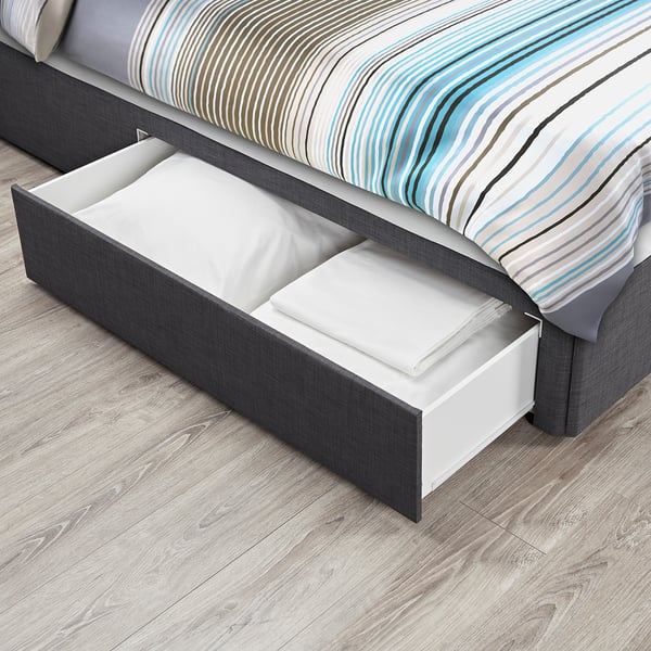 Bed with pull-out drawer for storage. Drawer is open, showing folded linens. Grey frame, patterned bedding.