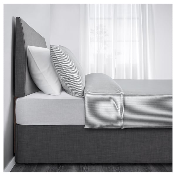 A bed with a grey, upholstered headboard and a striped mattress cover. It includes drawer storage under the bed. Curtains hang in the background.
