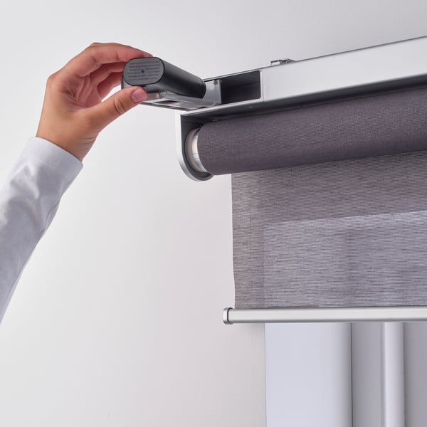 A hand holding a small black device connects it to a motorised roller blind, demonstrating its use to transfer back-up power.