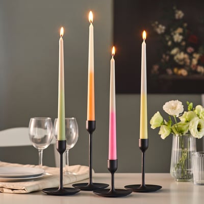Four colourful taper candles in black holders on a dining table, lit and creating a warm atmosphere for a dinner setting.