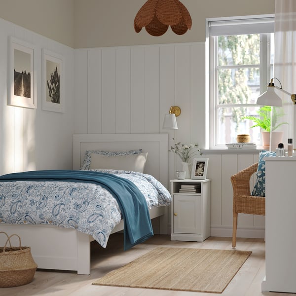 Cosy bedroom with natural light, white bed, blue blanket, small bedside table, books, and rattan chair.