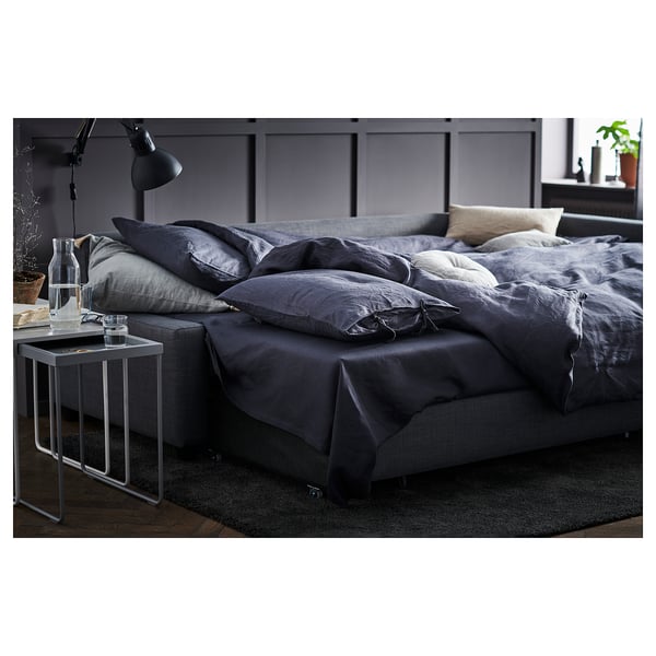 Cosy grey convertible sofa-bed with dark linens, black lamp, glass table, and plants.