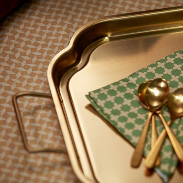 Gold-colored metal tray with handles on a patterned tablecloth, accompanied by matching patterned napkin and gold utensils.
