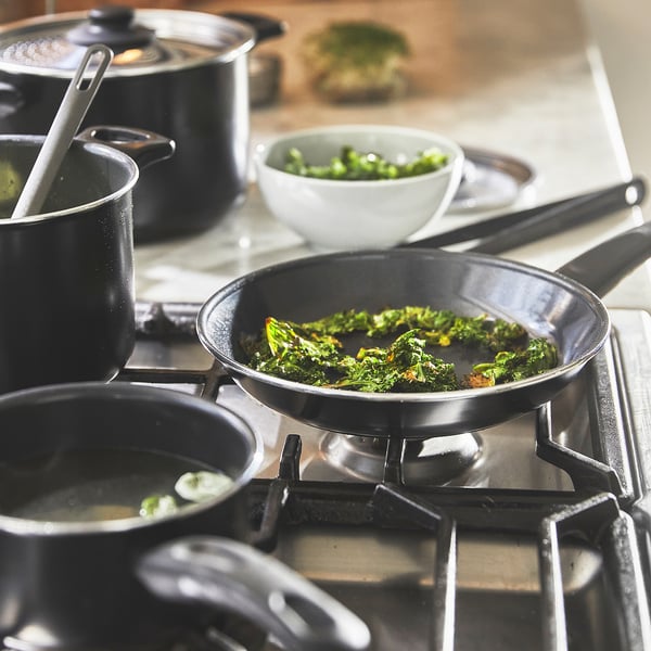 Cooking scene: pans on stove with greens. Even heat distribution shown. Comfortable handles present.