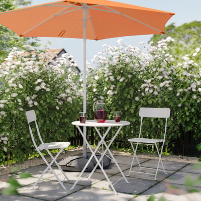 Patio with table, chairs, parasol, carafe, glasses, bushes, house.