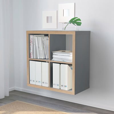 Grey KALLAX shelf with books, binders, plant, and empty frames in minimalist room.