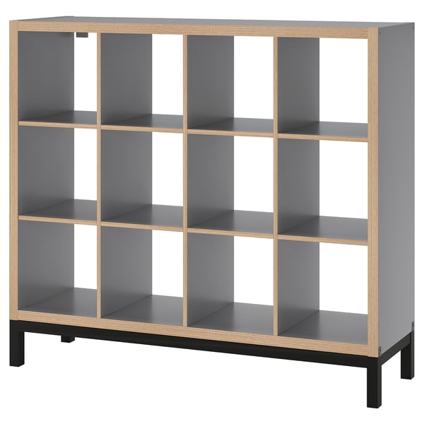 A KALLAX shelf unit in grey with light wood accents and black legs. It has a modular, cube-like design with open compartments.