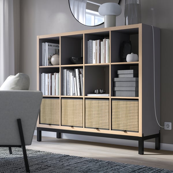 KALLAX grey bookshelf with open & closed storage, supported by black metal frame, holding books, decor, & boxes, positioned near a grey chair.