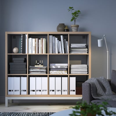 KALLAX light wood shelf, filled with books & decor. Tall, neatly organised, grey couch right, plant in front.