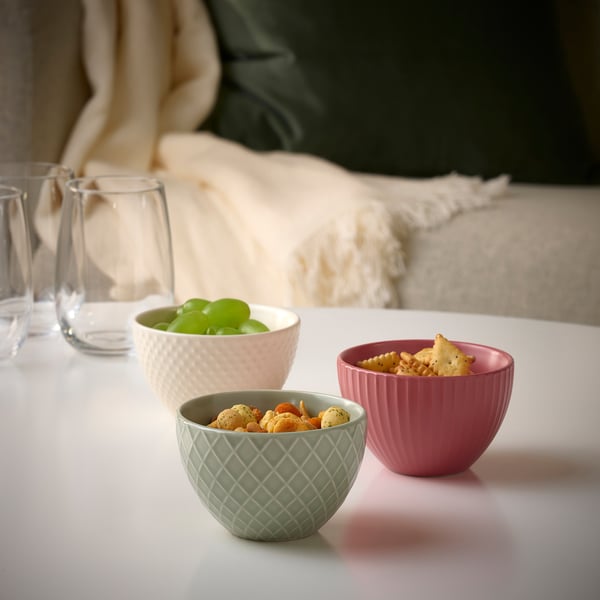 Three colourful bowls holding grapes, crackers, and snacks sit on a table, accompanied by clear glasses, suggesting a casual snack setup.