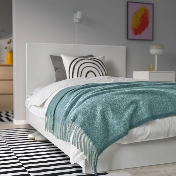 Modern bedroom with white bed, grey-turquoise throw, striped rug, art, lamp. Blanket is recycled polyester and stylish.