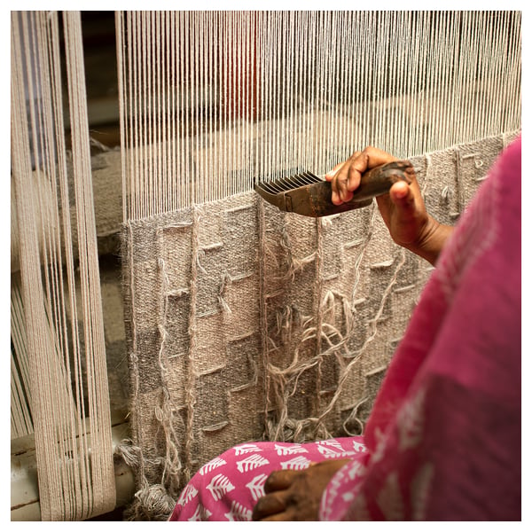 Person weaving rug using loom, holding tools, wearing pink. Traditional craft, handmade.