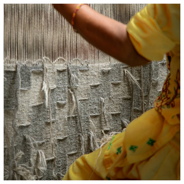Person weaving grey wool rug on loom, wearing bright yellow shirt.