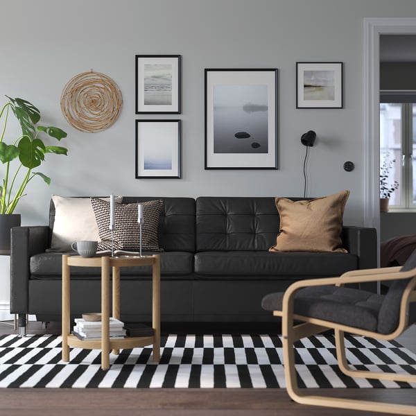 Modern living room with black leather landskröna sofa, patterned rug, wooden side table, and wall art. Sofa is large, central, with cushions.