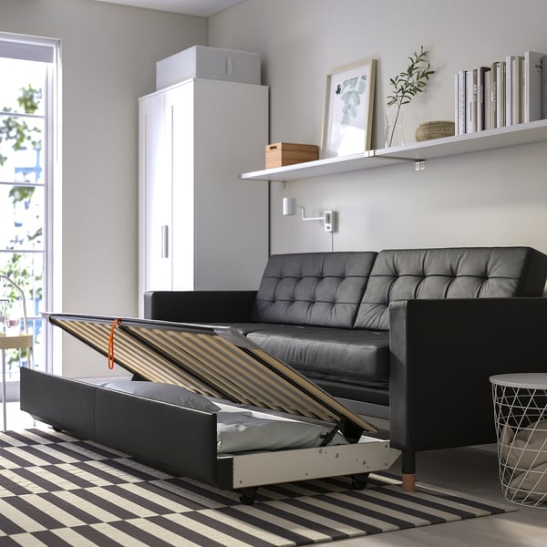 Modern living room with a black leather convertible sofa-bed, storage drawers open, near a shelf with books and a plant.