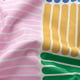 Bright, colourful, patterned fabric with overlapping stripes in pink, green, mustard, and blue hues. High-quality design, softly textured.