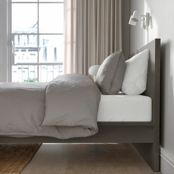 Modern bed with grey bedding and pillows, high headboard, positioned by large window with city view. Quality design and comfortable setup.