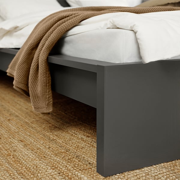 Bed frame close-up, clean modern design. White bedding, warm beige blanket, dark grey frame, natural textured rug.