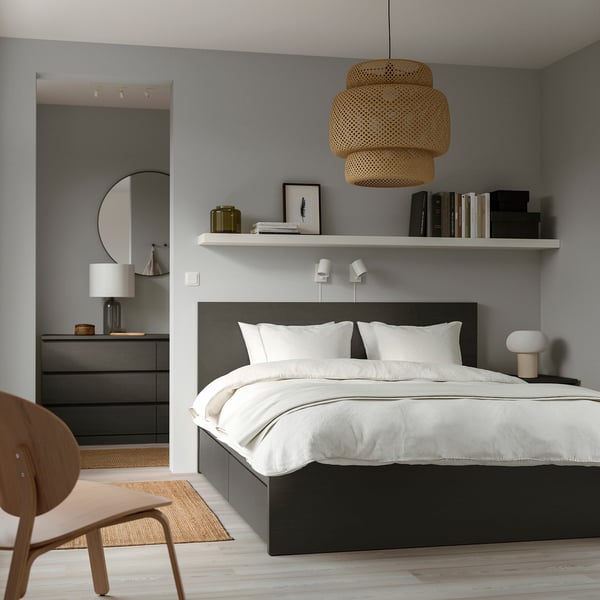 Minimalist bedroom with black MALM bed, crisp linens, wicker light, wooden chair, dresser, artwork, books on floating shelf, light floors, beige rug.