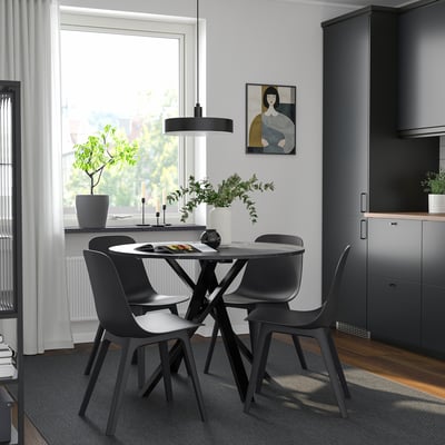 Modern dining area with MARIEDAMM black round table and chairs, sleek design, positioned near large window, greenery, and grey cabinets.