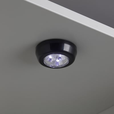 Small, round RAMSTA led light, black with clear cover, mounted on grey surface, emitting warm white light.