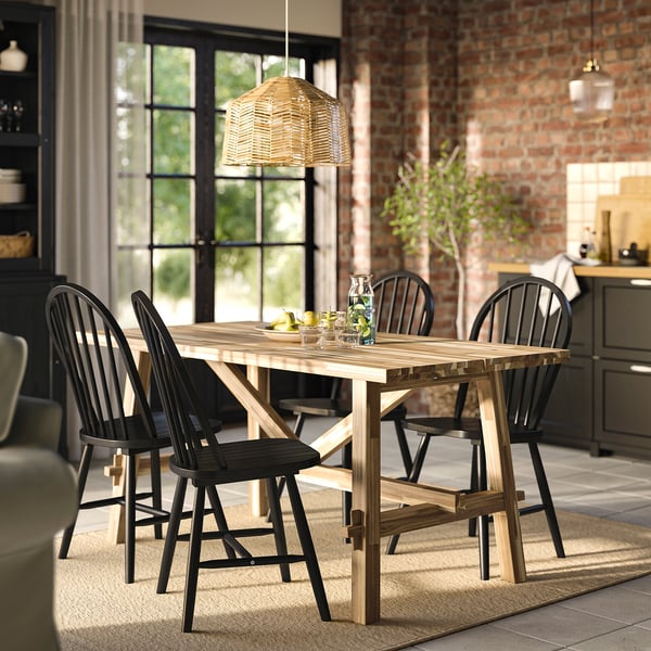 SKOGSTA dining table & chairs in warm acacia wood with pendant lamp, bowl of fruit, and glasses on rustic brick backdrop.