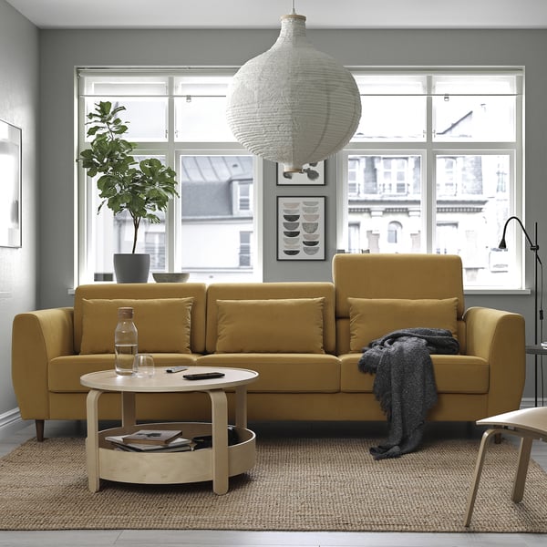 Modern living room with large yellow SLATORP sofa, round coffee table, grey rug, large plant, and hanging light. Large windows at the back.