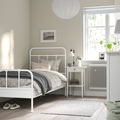 Minimalist bedroom STJÄRNÖ white metal bed, large window, cosy linens, nuitral decor. Matched bedside table, lamp, and plants.