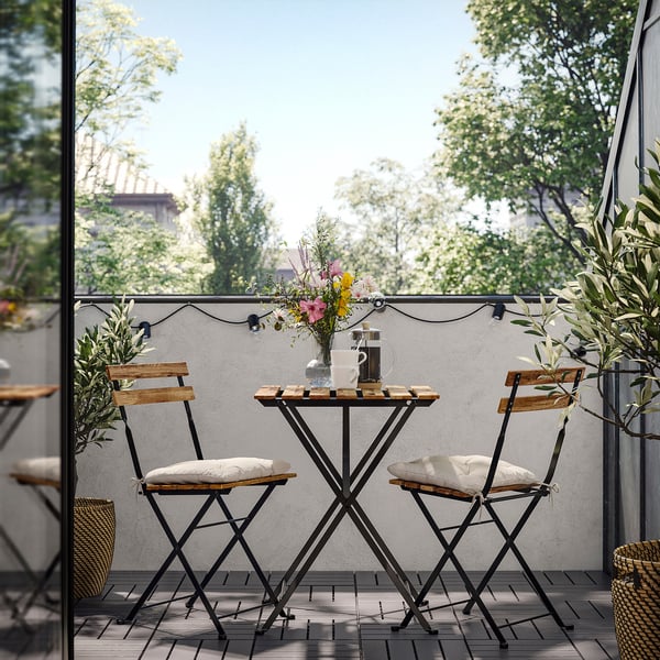 Small balcony setup with TÄRNÖ table and chairs. Wooden seats, black metal frames, outdoors. Fresh flowers on table, greenery around.