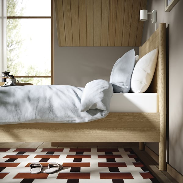 Bedroom scene with wooden bed, white sheets, three pillows, slippers on patterned rug, window with view.