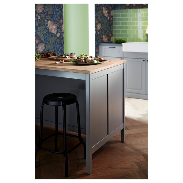 Modern kitchen island named TORNVIKEN in grey with wooden top, black stool, various vegetables, blue floral wallpaper, and green tiled backsplash.