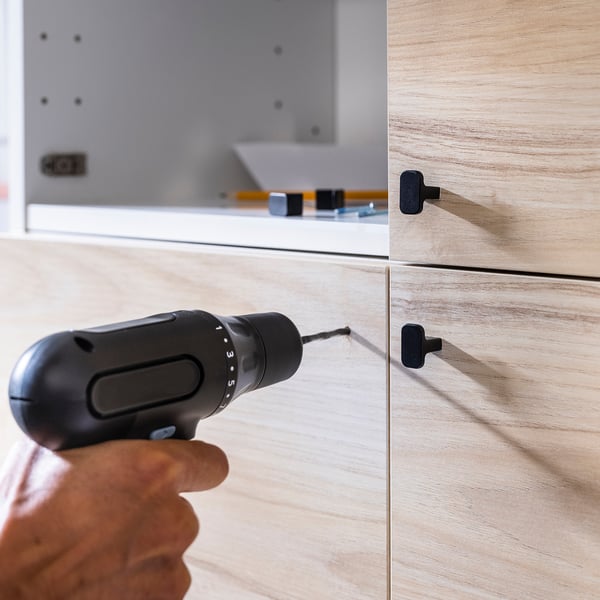 A hand holding a black TRIXIG cordless drill aiming at a wooden drawer. The drill has a rubber grip and an led light. The drawer black handles.