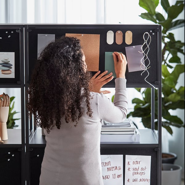 Person organising notes on a TROTTEN double-sided notice board and cabinet. The board divides the workspace, offering both open and locked storage.